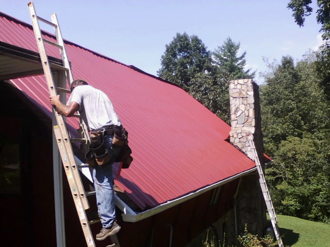 Roofer on a ladder beside a red metal roof and stone chimney — Soffit & Fascia Repair by Roofing Brothers in Mexia